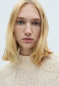 Geweven trui in lichtbeige met een gestructureerd patroon en geribde halslijn. Model heeft steil blond haar en een neutrale uitdrukking.