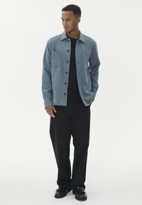 Man som bär en blå skjorta med knappar över en svart T-shirt, svarta byxor och svarta skor, står med en hand i fickan på vit bakgrund.