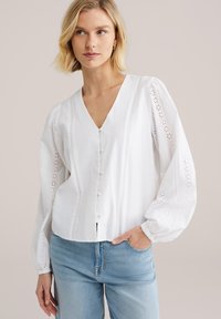 Blouse blanche boutonnée à col en V, manches bouffantes, détails décoratifs en œillets, et ourlet légèrement court, associée à un jean bleu clair.