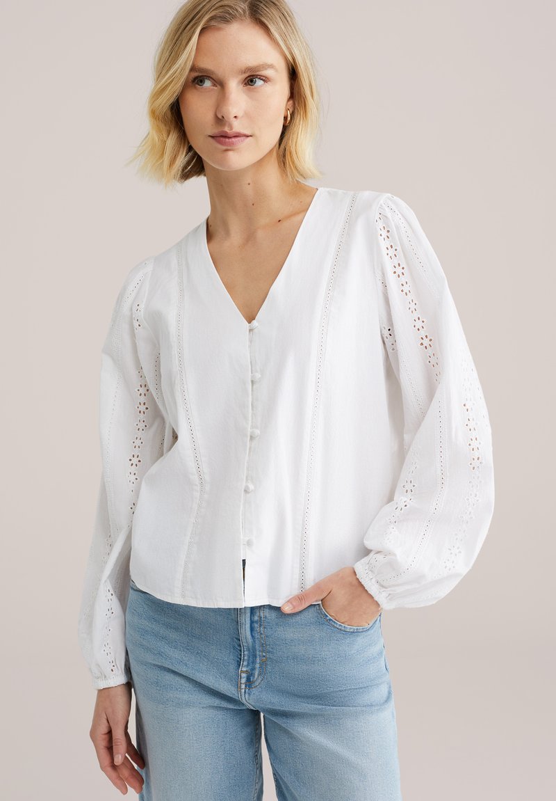 Blouse blanche boutonnée à col en V, manches bouffantes, détails décoratifs en œillets, et ourlet légèrement court, associée à un jean bleu clair.