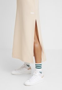 Béžová dlouhá sukně s postranním rozparkem. Nošena s bílými teniskami a zeleně pruhovanými ponožkami s logem listu. Hladká struktura látky.