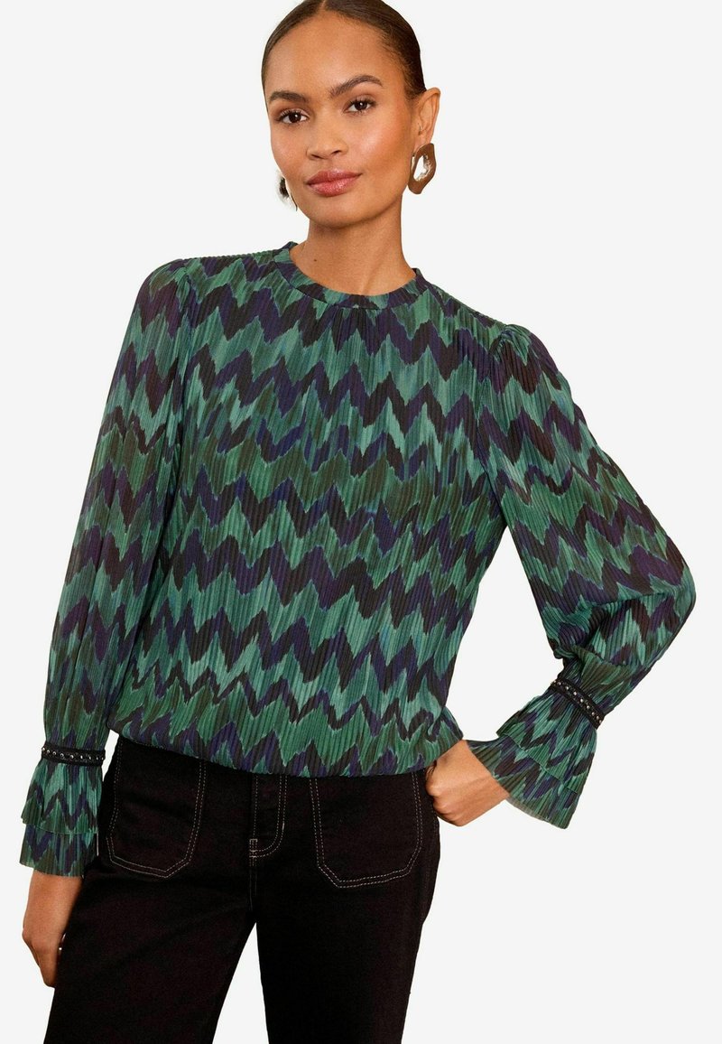 Blusa a maniche lunghe in verde e blu navy con motivo a zigzag, tessuto plissettato, scollatura rotonda, polsini arricciati con dettagli di perline. Indossata con pantaloni scuri.