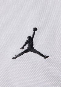 Sylwetka koszykarza skaczącego z rozstawionymi nogami, trzymającego piłkę jedną ręką, wyhaftowana na czarno na białej tkaninie.