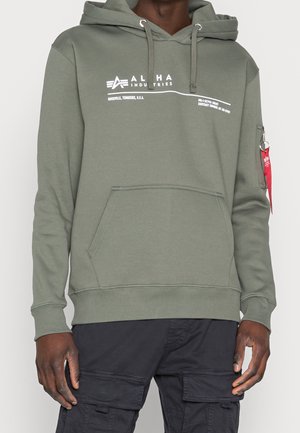 Oliwkowa bluza z kapturem z przednią kieszenią i białym logo Alpha Industries, noszona z czarnymi spodniami typu cargo i czerwoną metką na kieszeni na lewym rękawie.