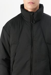Veste noire avec un col montant, fermeture zippée, tissu texturé et détails de couture en biais. Remarque : aucun matériel ou accent visible.