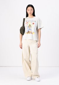 T-shirt gráfica cinza com um design floral de Matisse, combinada com calças brancas largas e ténis brancos. Acessórios incluem uma pequena bolsa verde oliva.