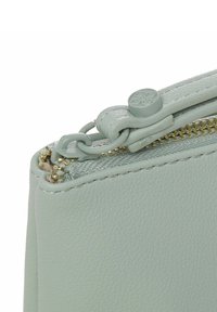Angolo di una borsa di pelle verde chiaro con zip dorata, superficie testurizzata e un tirazip rotondo. Cuciture visibili sulla parte superiore.