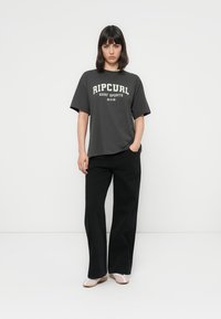 Ung kvinde står med den ene hånd i lommen iført en mørkegrå "Rip Curl Surf Sports" t-shirt, sorte vide bukser og hvide sko.