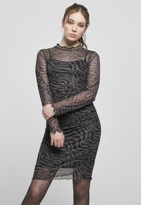 Permatomas juodas tinklinis suknelė su zebra raštu, ilgomis rankovėmis ir banguotu hemu. Prigludęs dizainas pabrėžia kūno siluetą.