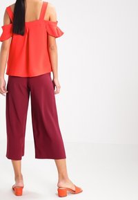 Červený off-shoulder top s volány, spárovaný s burgundskými širokými kalhotami. Modelka má na sobě oranžové sandály na blokovém podpatku.