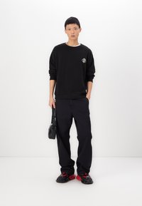 Zwarte sweatshirt met een grafisch ontwerp, gecombineerd met zwarte cargobroeken. Schoeisel heeft een rode accentkleur en er wordt een kleine zwarte tas meegenomen.