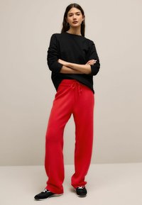 Zwarte longsleeve shirt gecombineerd met losse, felrode joggingbroek met een trekkoord in de taille, gedragen met zwarte sportschoenen.