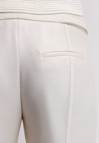 Close-up van beige stoffen broek met een achterzak en een textuurband met horizontale ribbels.