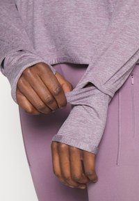 Langærmet, lyse lilla top med tekstureret stof. Har tommelhuller ved ærmekanterne og lagdelt design, kombineret med matchende leggings.