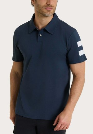 Uomo che indossa una polo blu navy a maniche corte con colletto e due strisce bianche sulla manica sinistra, in piedi davanti a uno sfondo semplice.