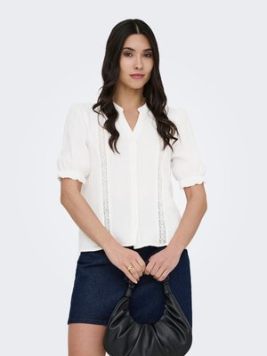 Kvinde med langt mørkt hår iført hvid bluse med pufærmer og mørkt skørt, der holder en lille sort håndtaske foran en lys baggrund.