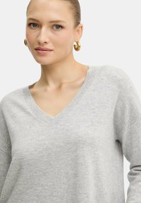 Donna con occhi azzurri che indossa orecchini a cerchio dorati e un maglione grigio chiaro a V, su uno sfondo bianco uniforme.