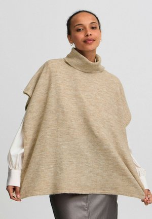 Poncho en tricot beige avec un col montant. Présente une coupe décontractée et une texture douce, superposé à une chemise blanche à manches longues.