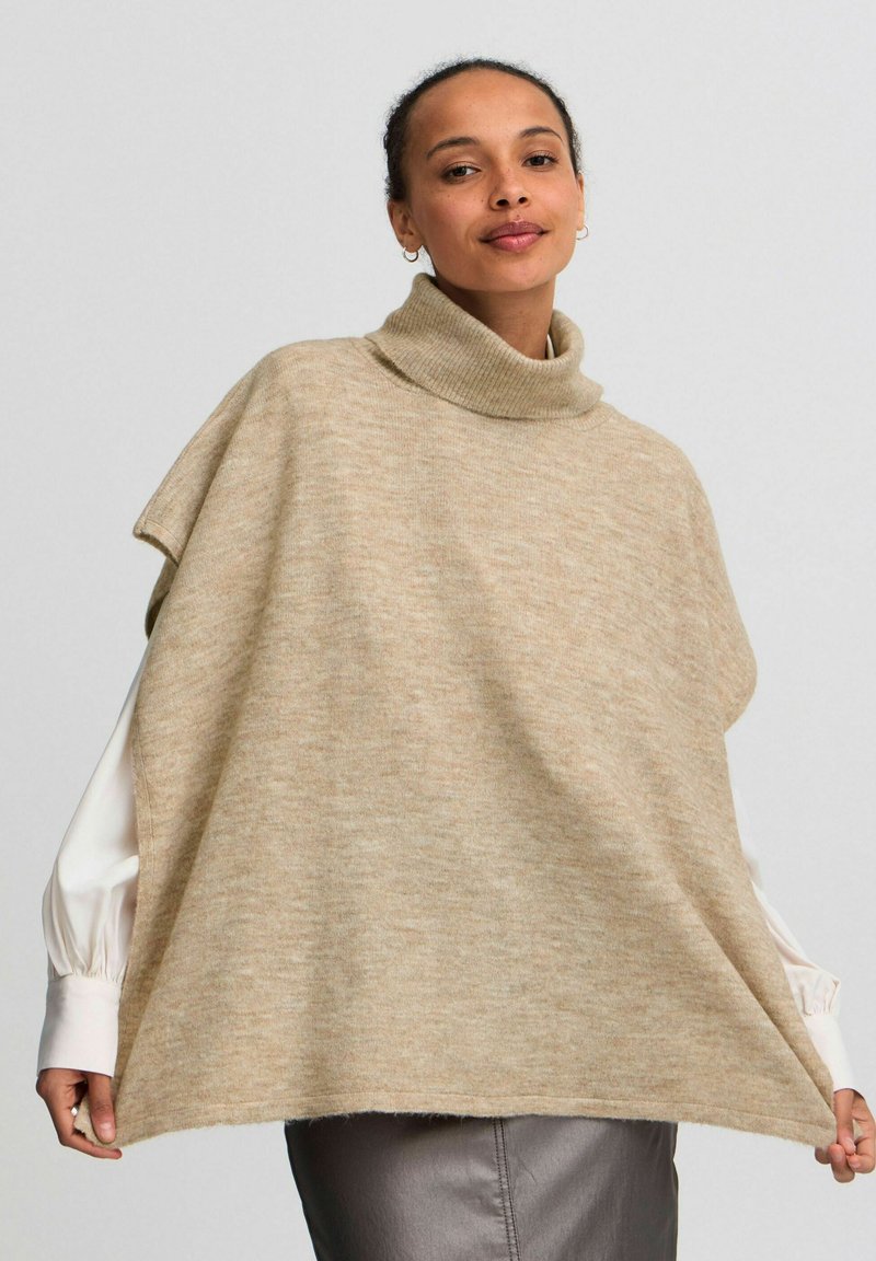 Beige, gebreide poncho met een hoge kraag. Kenmerkt zich door een ontspannen pasvorm en zachte textuur, gelaagd over een wit overhemd met lange mouwen.