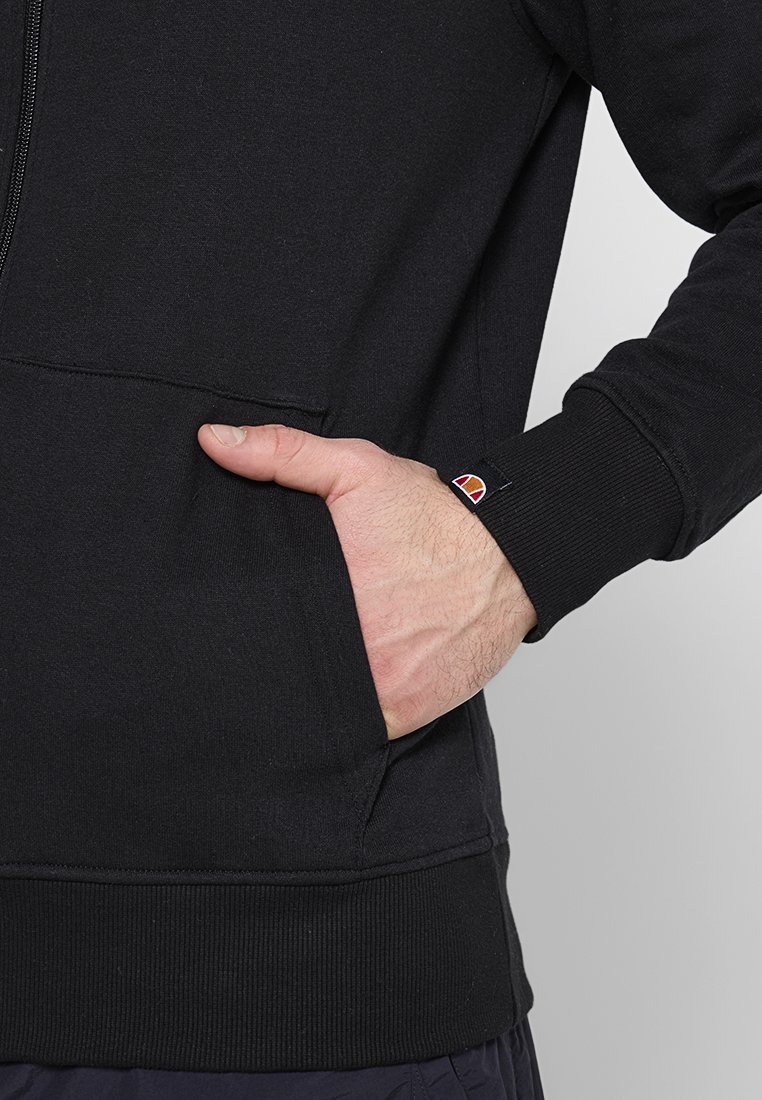 Mano en el bolsillo de una sudadera con cremallera negra con un pequeño logo rojo, naranja y blanco en el puño de la manga sobre un fondo liso.