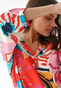 Farverig, mønstret bluse med store blomstermotiver i rød, pink, blå og gul. Blødt stof med v-hals og knaplukning ved ærmerne.