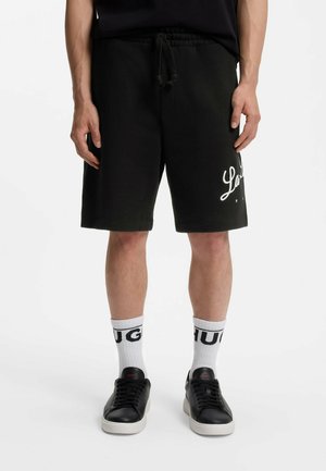 Uomo che indossa pantaloncini neri con scritta bianca, calzini bianchi crew con logo HUGO nero e sneakers basse nere con suola bianca.