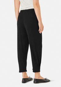 Zwarte, lichte, elastische broek met een tapered pijp en gathered boorden. Gedragen met zwarte flats, weergegeven vanaf de achterkant.