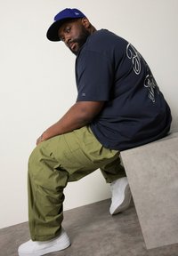Marineblå t-shirt med hvidt skrift, oliven cargo-bukser og hvide sneakers. Modellen poserer siddende på en grå geometrisk platform.