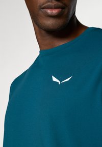 Τίρσινα αθλητική μπλούζα σε χρώμα τυρκουάζ με κανονική λαιμόκοψη, κοντά μανίκια και λευκό λογότυπο στο στήθος. Το ύφασμα φα ίνεται λείο και ελαφρύ.