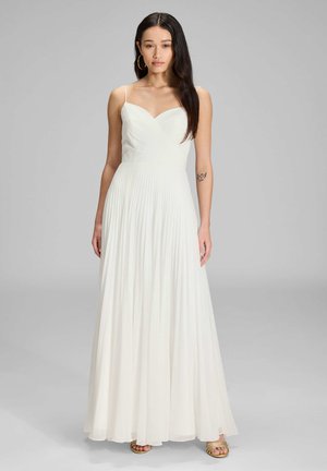 Robe longue plissée blanche avec un décolleté en cœur et des fines bretelles, mettant en valeur un corsage lisse et une jupe fluide, accompagnée de sandales dorées.
