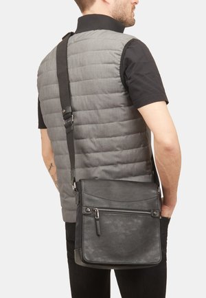Black leather messenger bag with a textured finish, featuring a front zip pocket and adjustable strap, worn crossbody over a gray puffer vest.