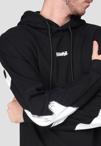 Uomo che indossa un hoodie nero con pennellate astratte bianche sulle maniche e un piccolo logo bianco sul petto, braccia incrociate.