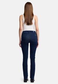 Jeans ajustados de mezclilla oscura, con una cinturilla estándar y dos bolsillos traseros, llevados con una camiseta sin mangas blanca y zapatos negros.