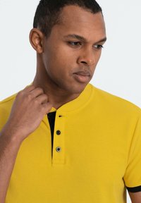 Gul polo skjorte med et tekstureret stof, der har en sort knapstolpe, tre knapper og en krave. Nærbillede af modellen.