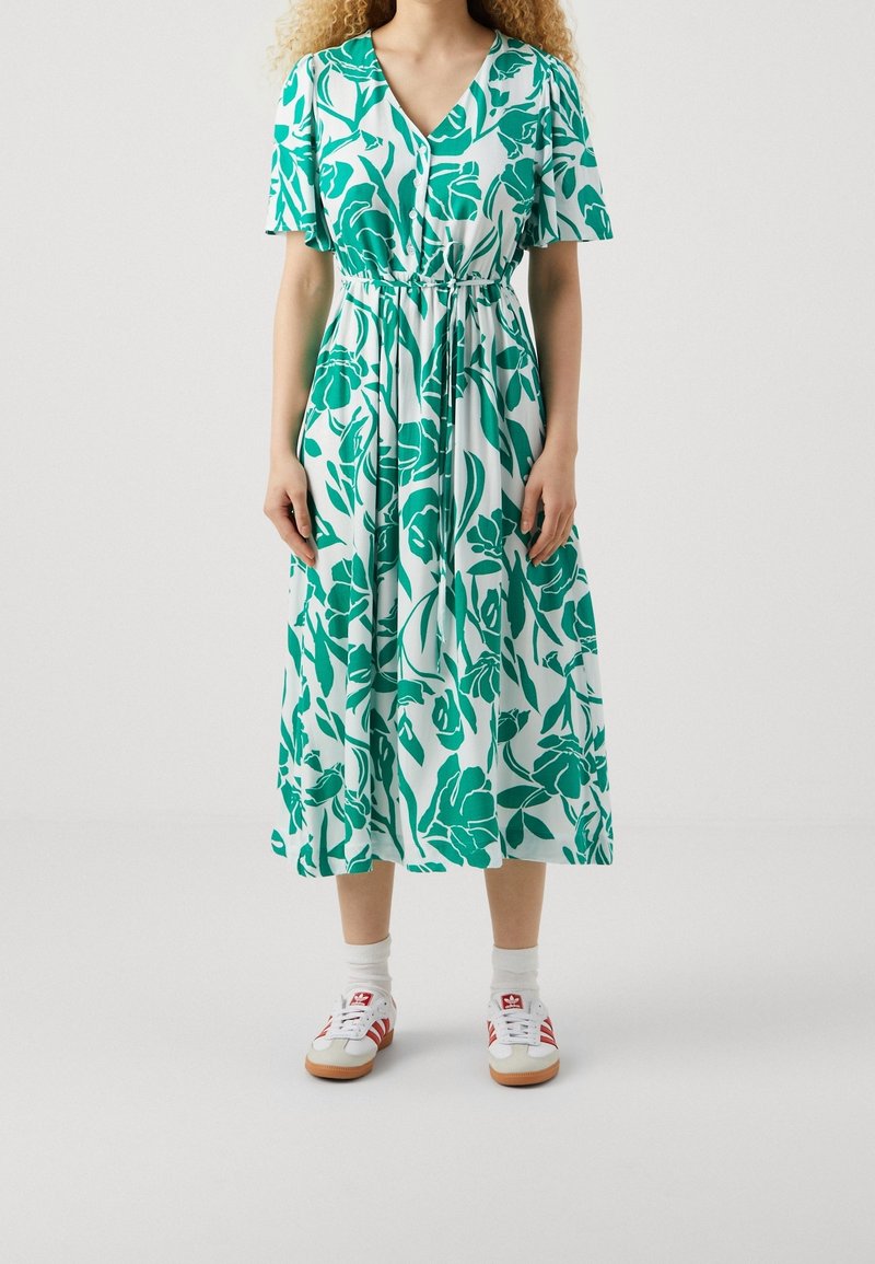 Femme portant une robe midi blanche à imprimé floral vert, à manches courtes, avec des boutons sur le devant et des baskets blanches à rayures rouges.