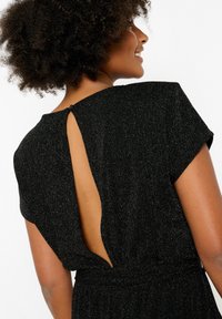 Melns kombinezons ar īsām piedurknēm, teksturēta auduma, kas izceļas ar spīdīgu apdari, un atslēgas cauruma aizmugures detaļa, ko nostiprina pogas.