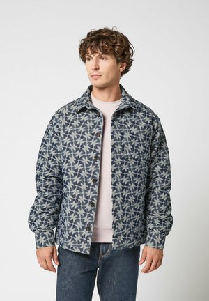 Jeune homme aux cheveux bouclés portant une veste boutonnée bleue à motifs par-dessus une chemise rose pâle et un jean bleu, regardant sur le côté.