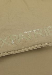 Hnedá látka s hladkým povrchom obsahuje slovo "EXPATRIET" vo veľkých, lesklých písmenách. Okraj je zošitý kontrastnou niťou.