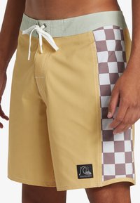 Bañadores de hombre en amarillo con una cinturilla verde claro. Presentan un patrón de tablero de ajedrez en marrón y blanco en el lateral y un cordón blanco.