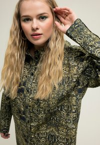 Camicia a stampa serpente verde e nera con apertura a bottoni sul davanti, maniche lunghe e vestibilità rilassata. Presenta un tessuto testurizzato e uno stile oversize.