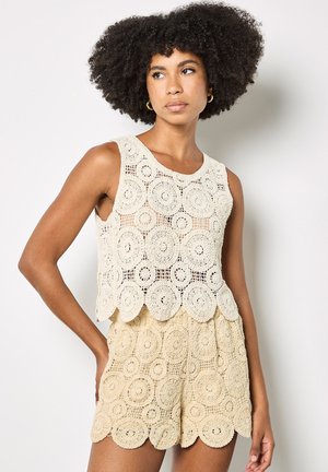 Frau mit natürlichen lockigen Haaren, die ein ärmelloses beiges Häkeloberteil und passende Shorts mit kreisförmigen Mustern trägt, stehend vor einer schlichten Wand.