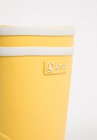 Botte en caoutchouc jaune à texture lisse, avec une bande blanche en haut et un logo en relief sur le côté.