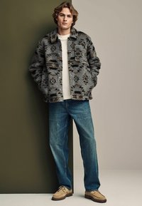 Fuzzy gray jacket with brown accents, a button front, and two pockets, paired with light blue jeans and beige shoes. Simple backdrop.