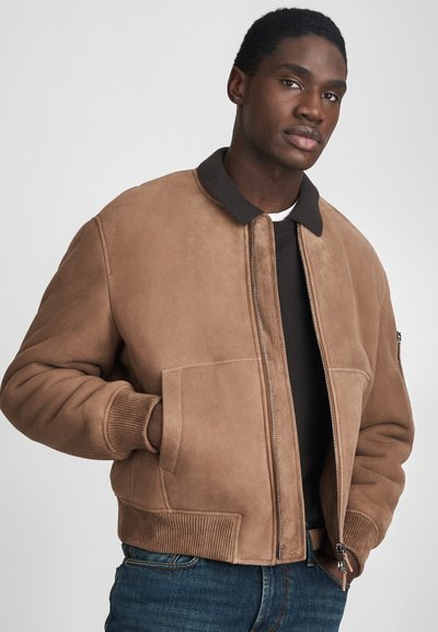 uomo che indossa un bomber in suede beige con un colletto e polsini scuri, una camicia scura e jeans blu, con le mani nelle tasche della giacca, rivolto in avanti.