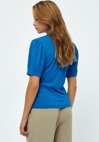 Mėlyna nėriniuota trumpomis rankovėmis palaidinė su prigludusiu dizainu ir subtiliomis pūstomis rankovėmis, derinama su šviesiais beige kelnėmis. Lygi tekstūra, vientisa spalva.