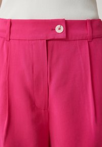 Pantaloni a vita alta rosa brillante con chiusura a bottone bianco e parte frontale plissettata, indossati con una maglietta bianca a costine.