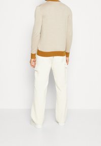 Pull en tricot beige avec un col et un ourlet côtelés marron, assorti à un pantalon cargo blanc cassé et des baskets blanches. Design minimaliste, texture douce.