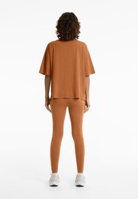 Túlnőtt narancssárga pamut póló rövid ujjakkal, testre simuló narancssárga leggingssel párosítva. A modell világos sportcipőt visel. Egyszerű háttér.
