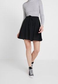 Person som bär en svart kjol med knytmidja, randig långärmad tröja och svarta högskor som går mot en enfärgad bakgrund.