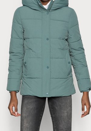 Persona che indossa una giacca imbottita con cappuccio verde acqua con chiusura a scatto e tasche laterali, abbinata a jeans neri sbiaditi.